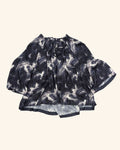 A short-sleeve blouse with a black and white abstract brushstroke pattern, featuring a gathered neckline with a bow detail, laid flat on a light cream background.