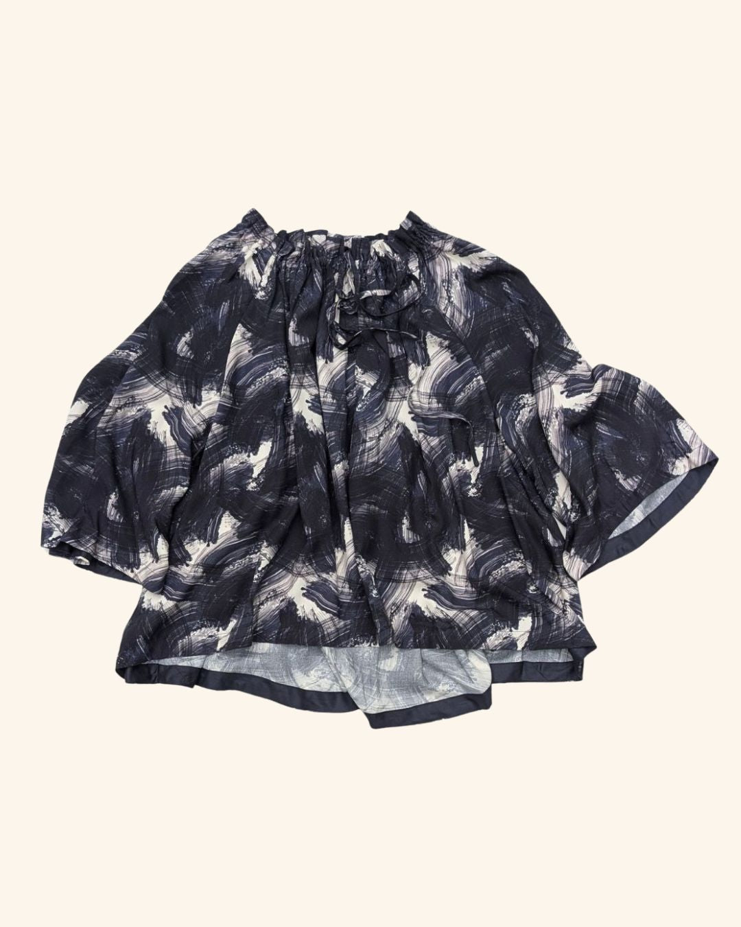 A short-sleeve blouse with a black and white abstract brushstroke pattern, featuring a gathered neckline with a bow detail, laid flat on a light cream background.