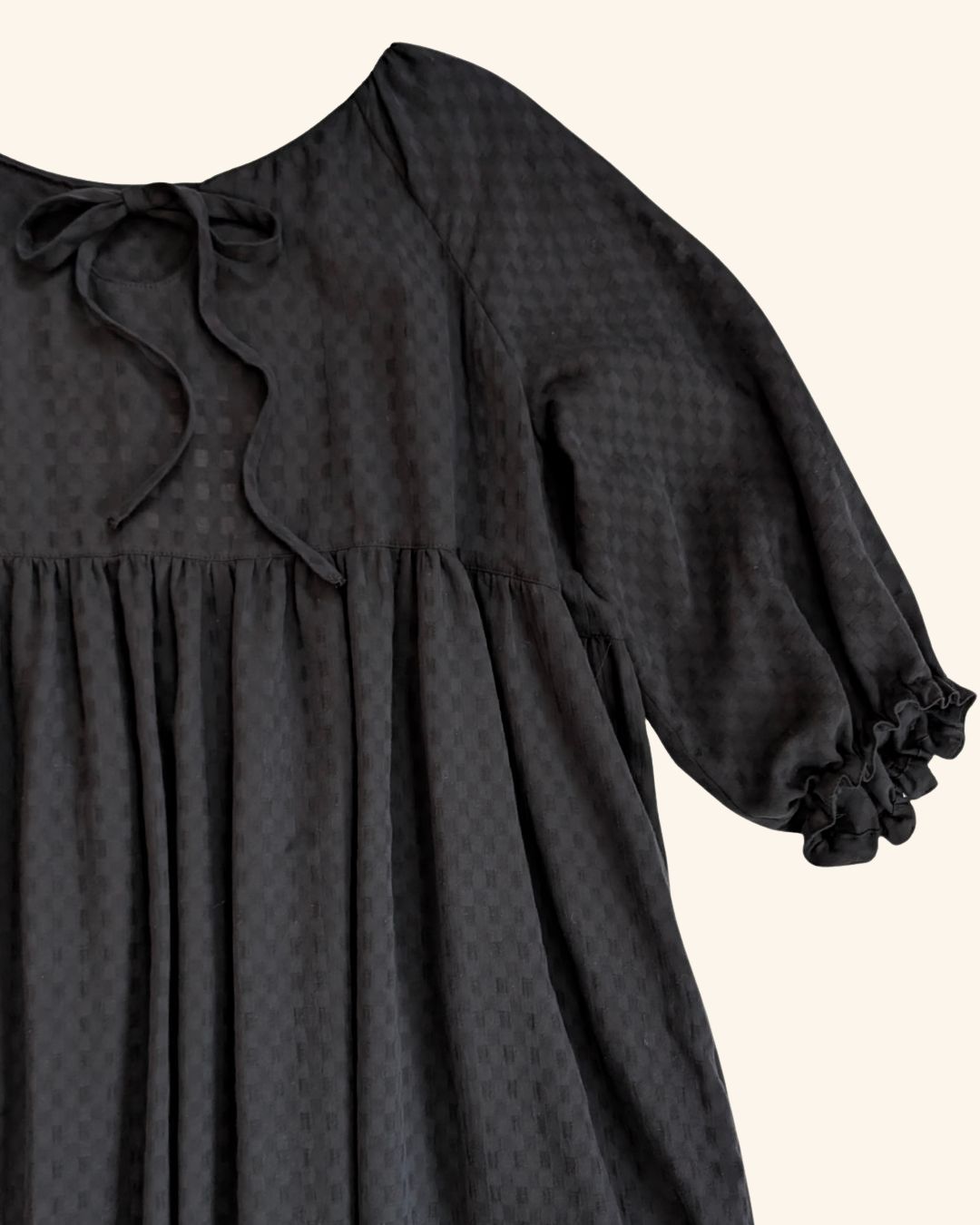 A close-up of a black textured dress with a subtle checkered pattern, featuring a tie-neck detail and ruffled elbow-length sleeves, set against a solid cream background.