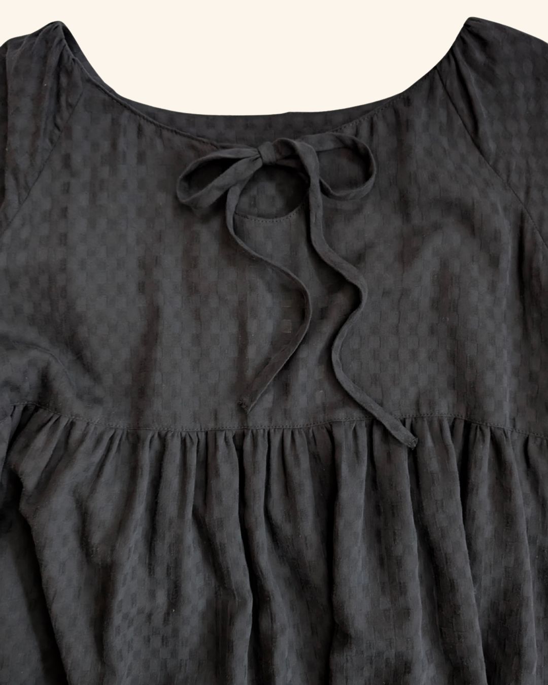 Close-up of a black textured dress with a tie-neck detail and gathered ruffle hem, shown against a light cream background.