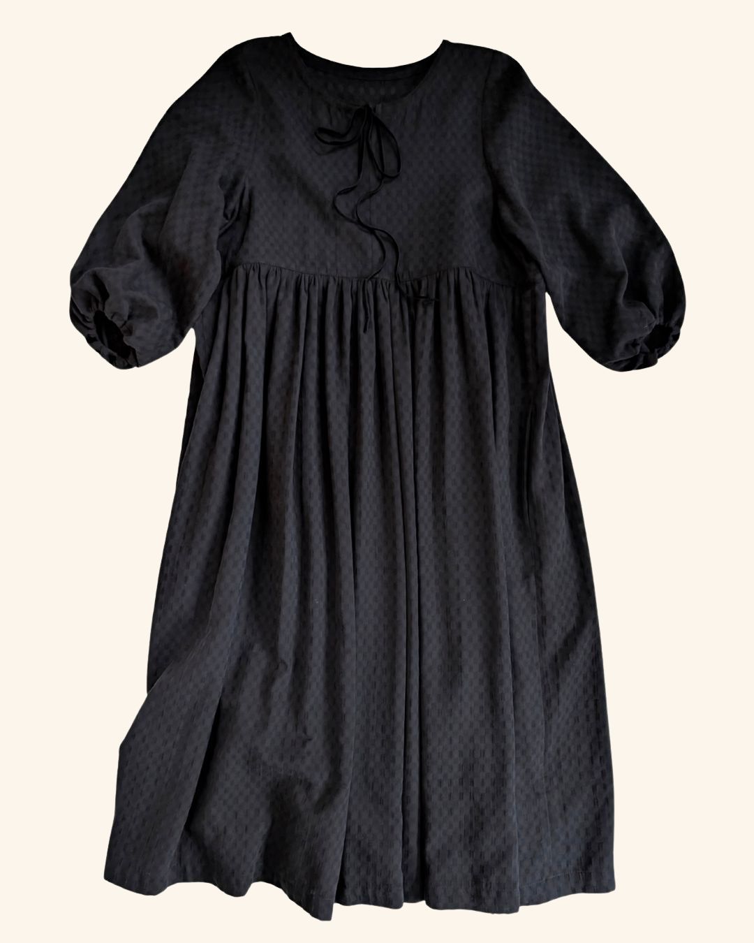 A black midi-length dress with a subtle repeating pattern, featuring 3/4 puff sleeves, a gathered waist, and a small bow detail at the neckline, shown against a solid cream background.